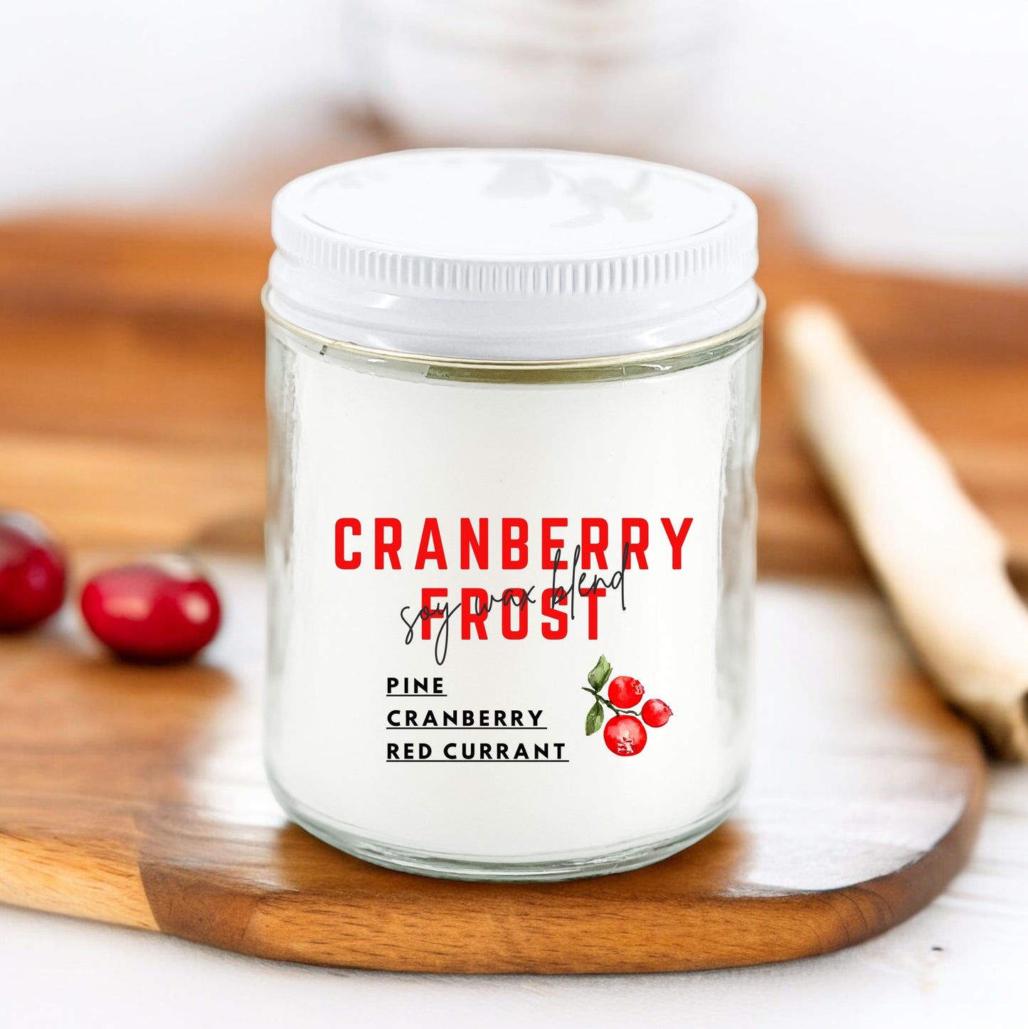 A white candle with the label 'CRANBERRY FROST' and additional text 'Pine Cranberry Red Currant' on it, placed on a wooden surface with cranberries and a cinnamon stick in the background.
