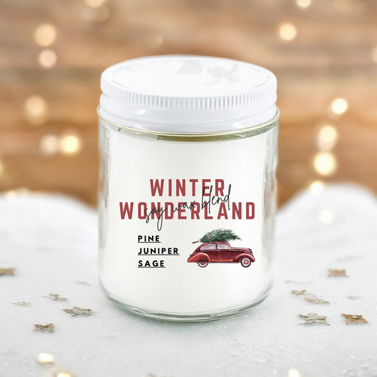 A white candle with a label reading 'Winter Wonderland', featuring illustrations of pine trees and a red car with a Christmas tree on its roof, set against a wooden background with lights and stars.