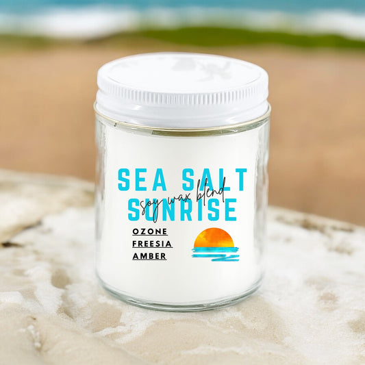 An 9 oz jar candle with the label 'Seasalt Sunrise' featuring a white background with blue and orange text, placed on a surface with a blurred background suggesting a beach setting.