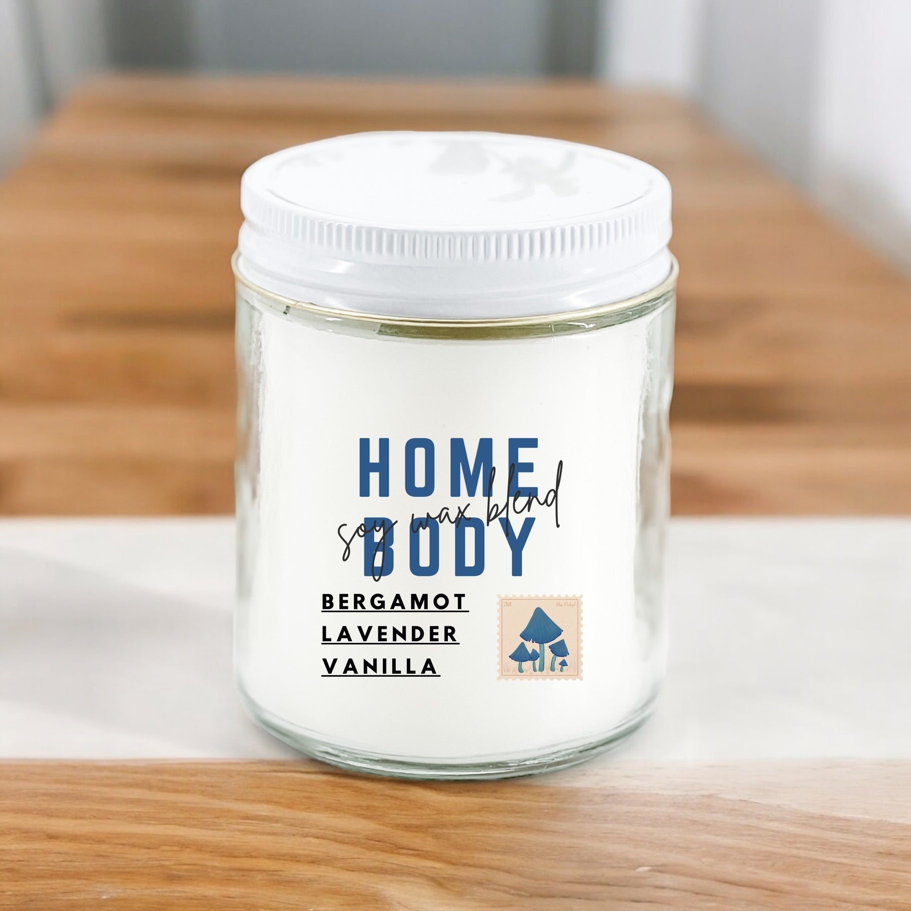 A white candle with the label 'Homebody' written on it, along with the scents 'Bergamot', 'Lavender', and 'Vanilla' listed below. The candle is placed on a wooden surface.